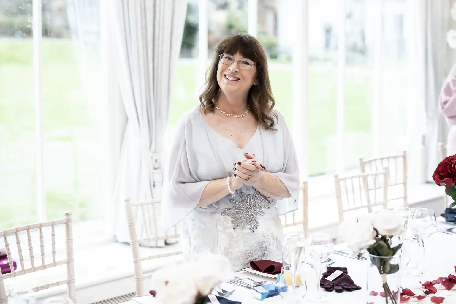 A smiling woman with long hair and glasses stands near a table set for an event, dressed in a light-colored outfit with floral detailing, in a bright room with large windows.