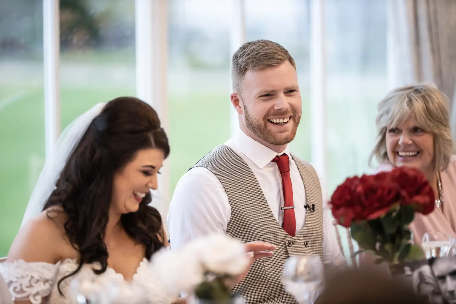 A bride and groom sitting and smiling during a wedding reception, with a woman beside them also smiling. There are flowers and glasses on the table in front of them.