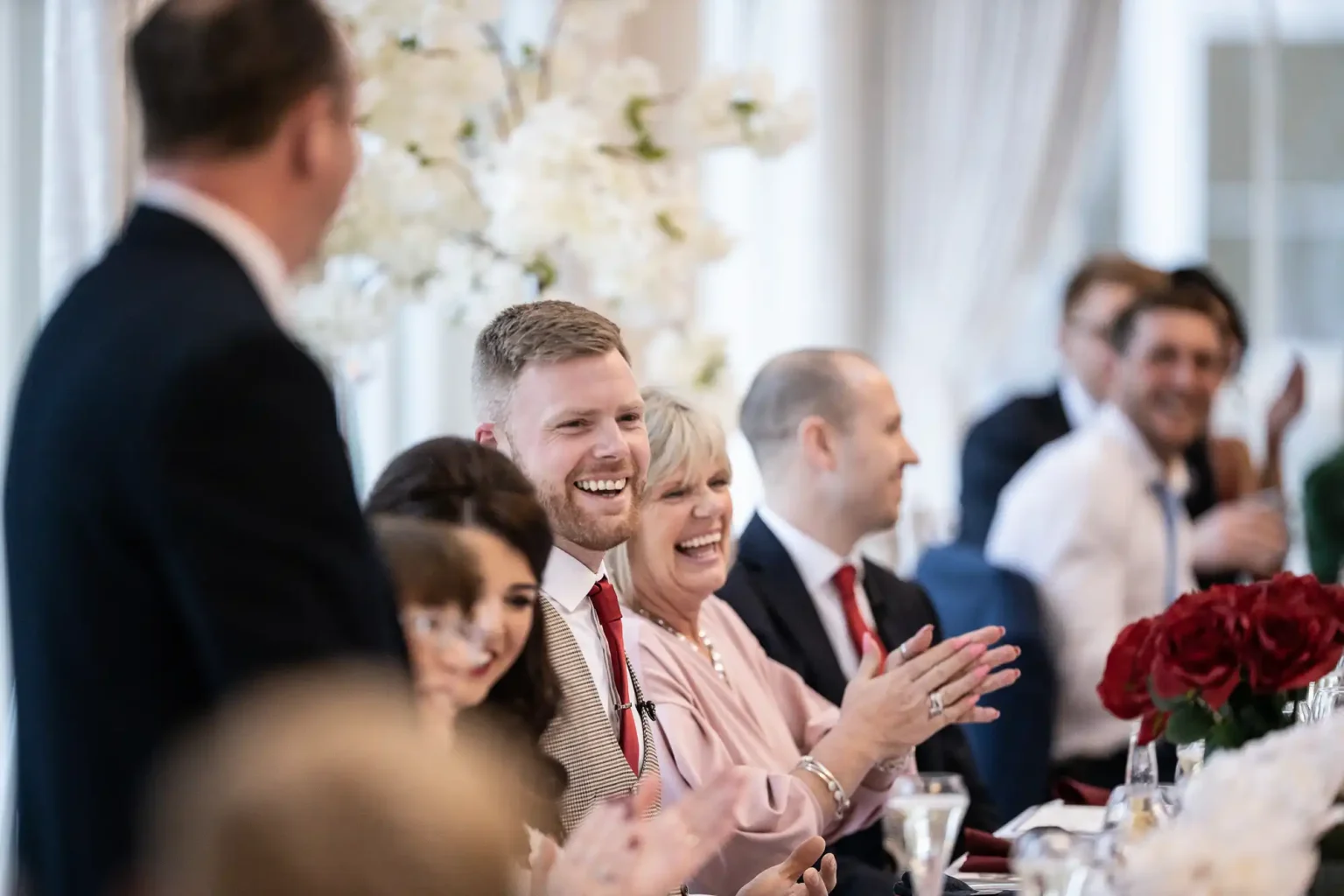 A group of people in formal wear are seated and clapping at an event. A man standing in the foreground appears to be speaking. White flowers and red roses are seen as decorations.
