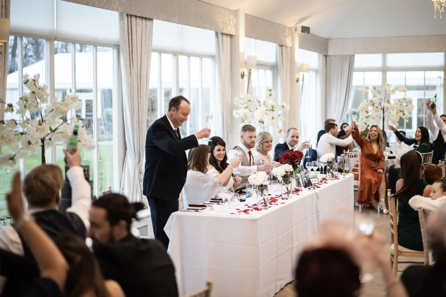 A group of people sitting at a long table in a well-lit event space, toasting with drinks. The table is decorated with flowers and candles.