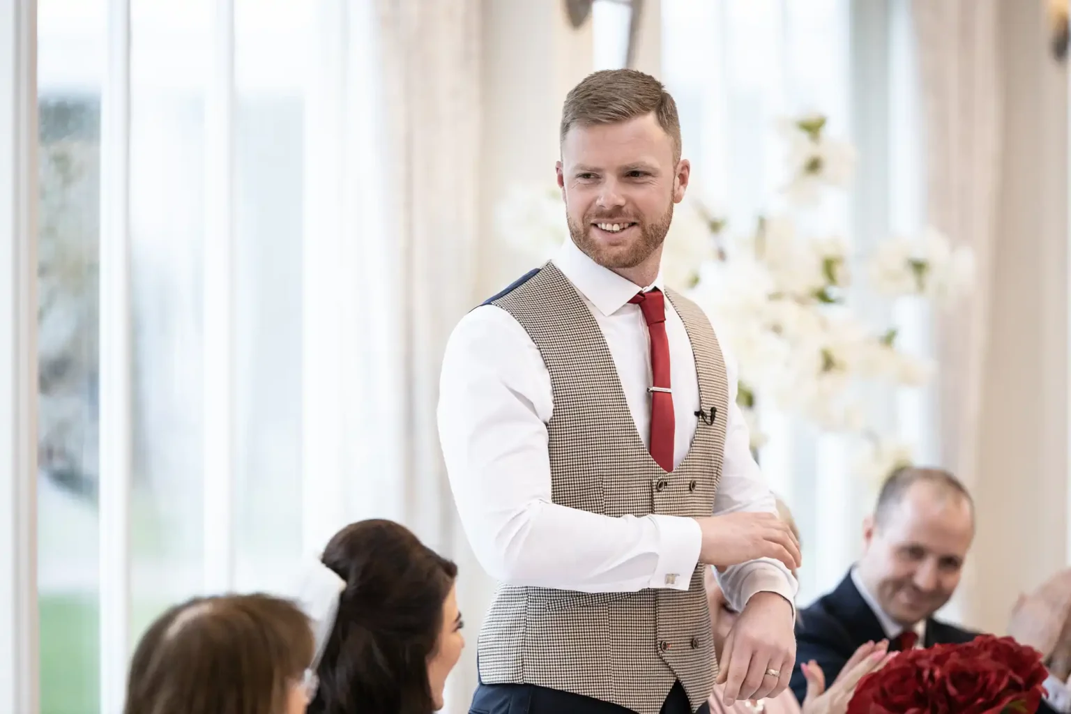 A man in a beige vest and red tie stands and speaks at an indoor event. People seated around him are listening attentively. White flowers are in the background.