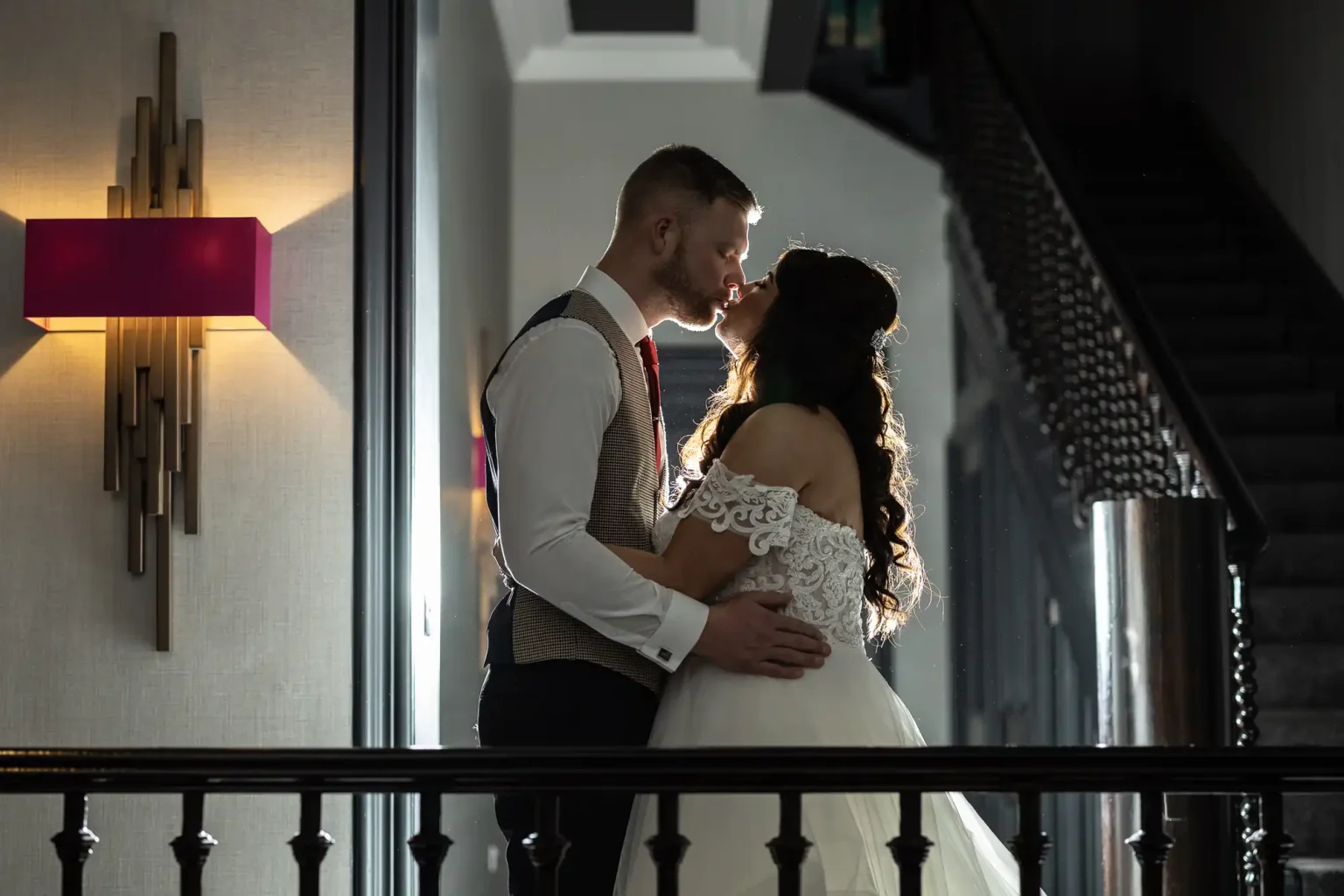 A couple in formal attire shares a kiss in a dimly lit indoor setting, with a staircase visible in the background. The woman is in a white dress and the man wears a vest and tie.