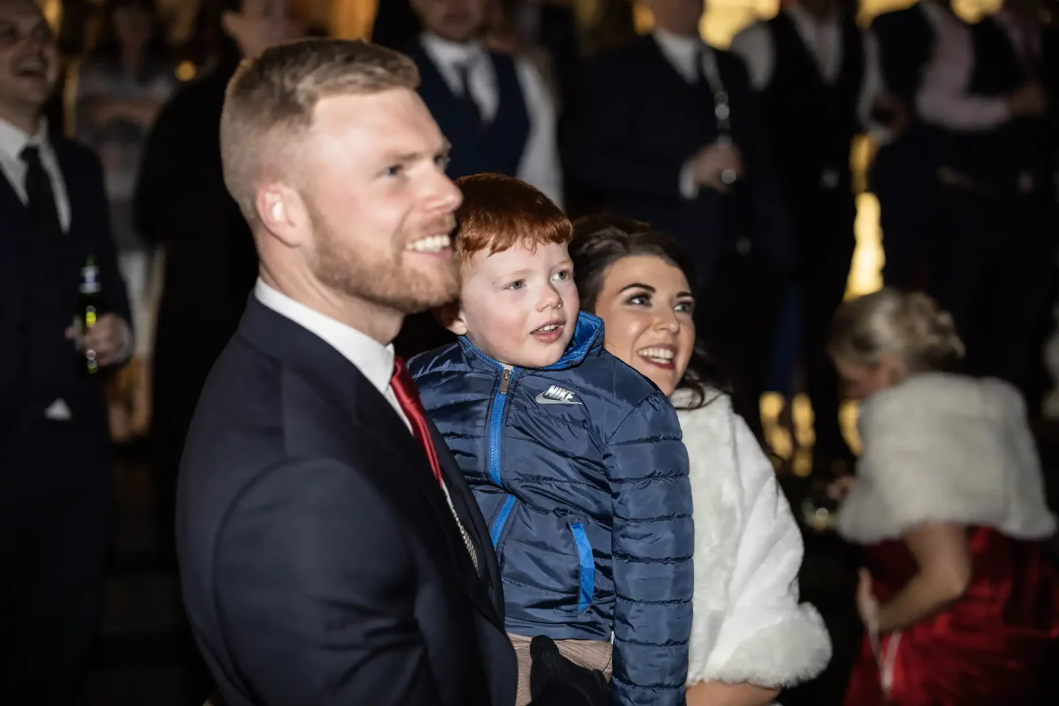 A man in a suit, a woman in a white outfit, and a child in a blue jacket stand together in a crowd, looking in the same direction.