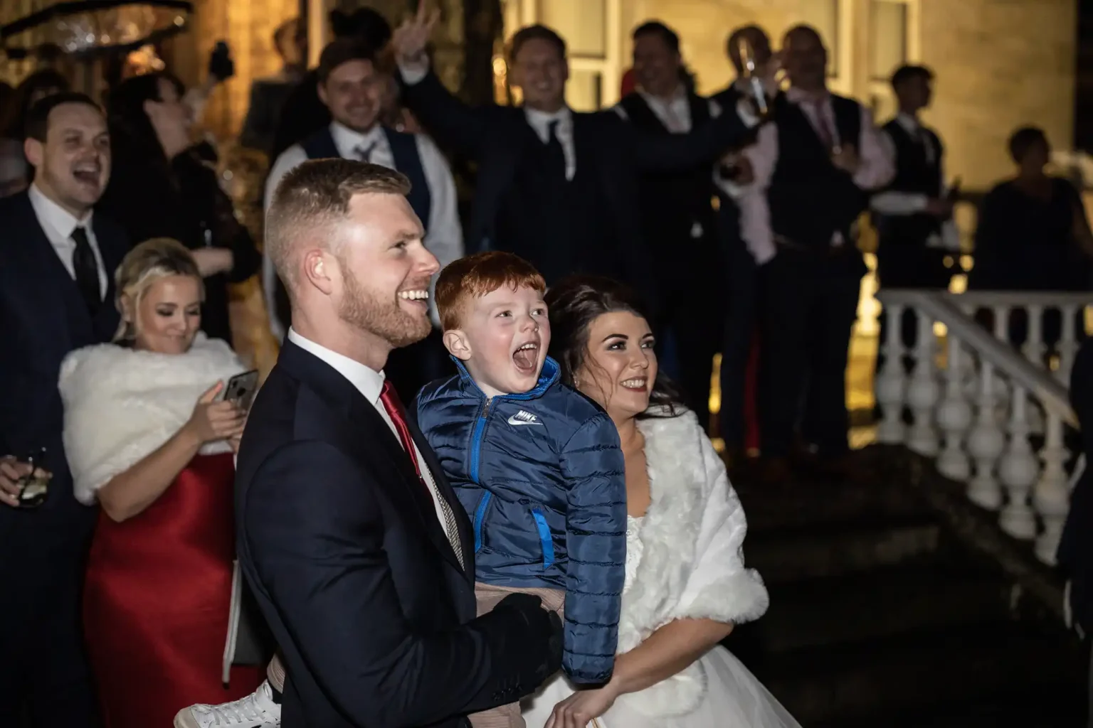 A smiling couple in formal attire holds an excited young boy at an outdoor evening event. People dressed in suits and dresses stand in the background, clapping and cheering.