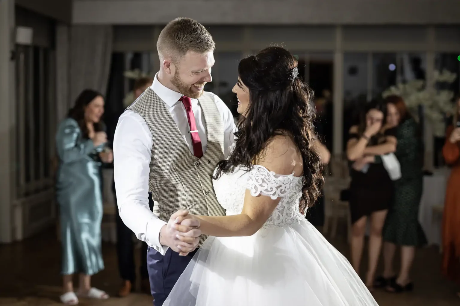 A bride and groom share a dance at their wedding reception while a group of guests watch and take photos in the background.