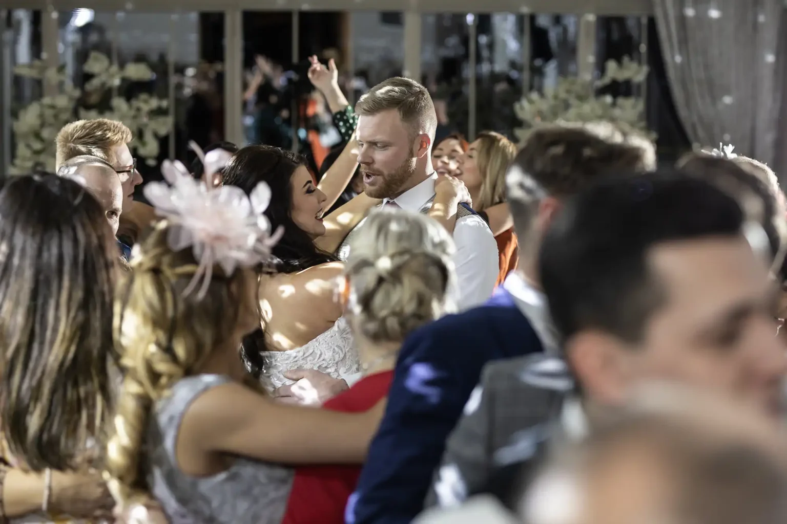 A couple dances in the center of a crowded ballroom, with others dancing around them. The room is decorated with flowers and drapes.