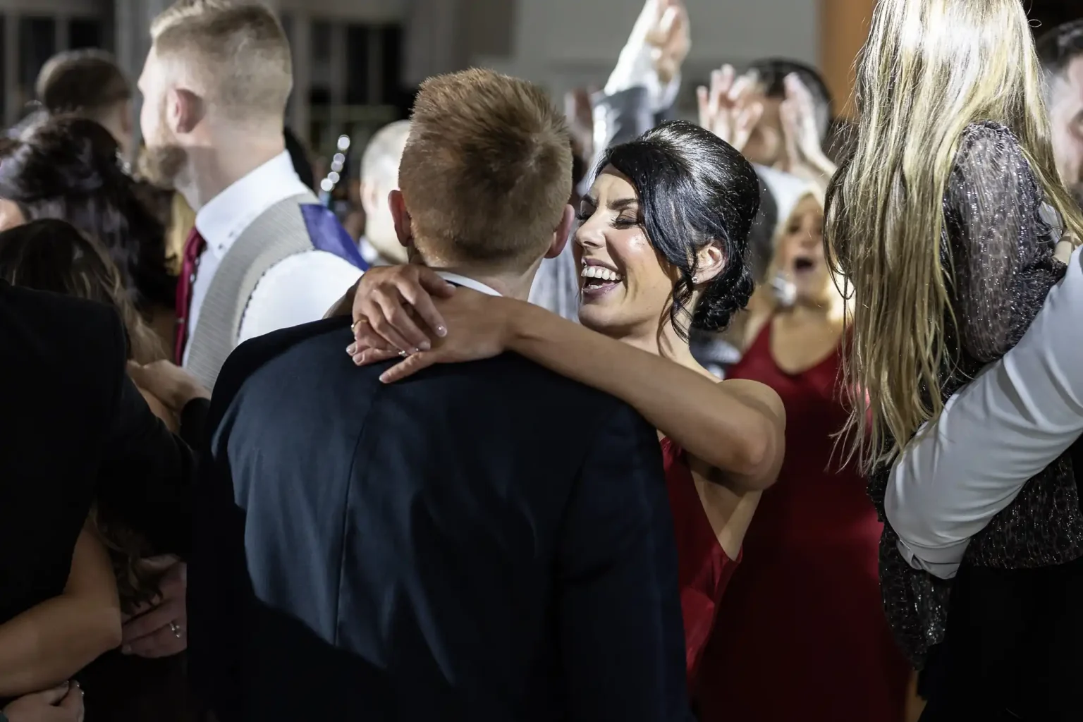 A woman in a red dress smiling and hugging a man in a dark suit at a crowded event, surrounded by other people dressed formally.