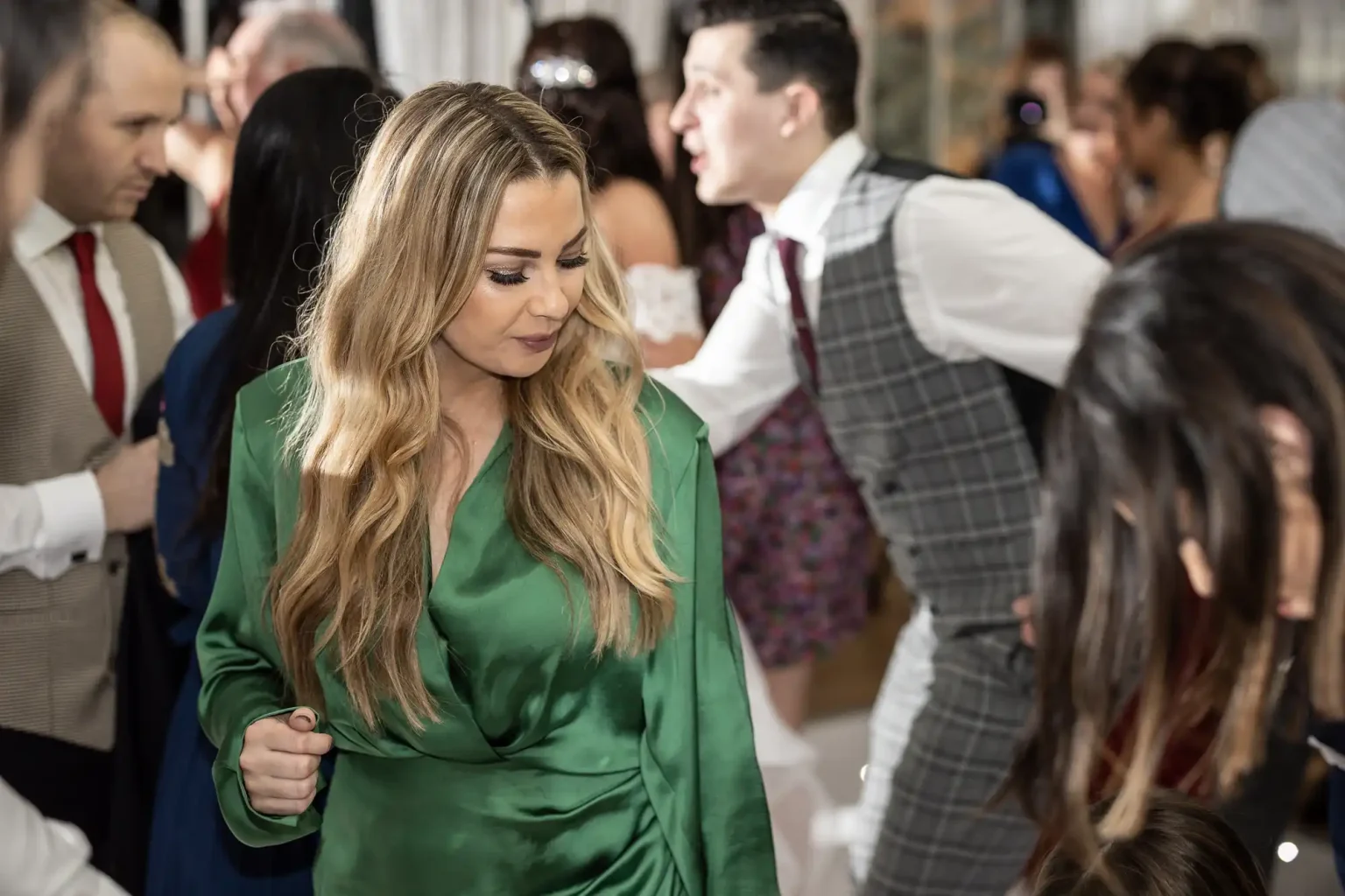 A group of people at a social event, with a woman in a green dress in the foreground and others in formal attire interacting in the background.