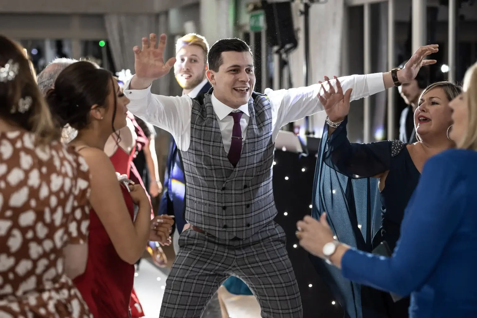 A man in a gray checkered suit dances enthusiastically with a group of people at a party.