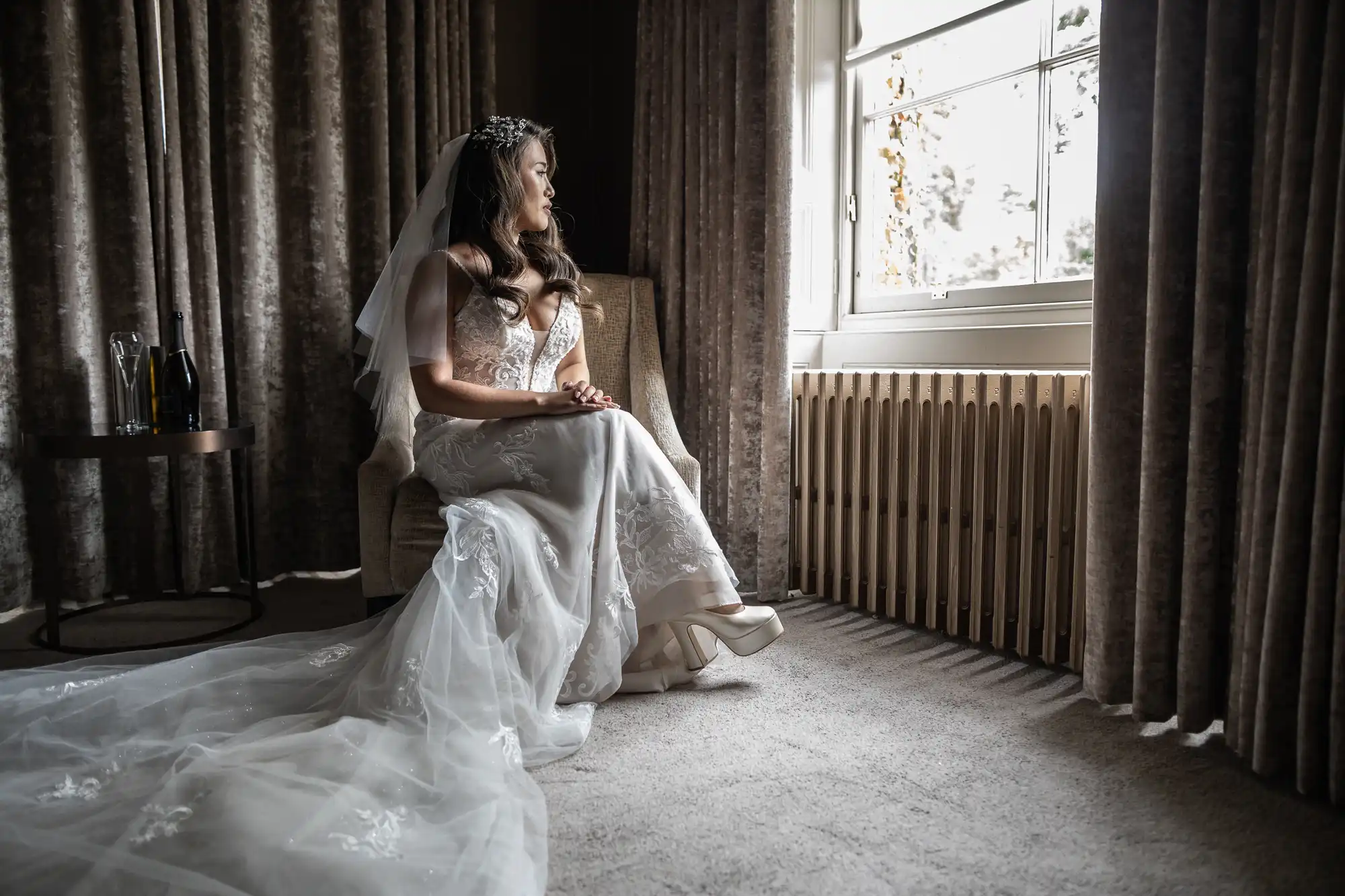 wedding photography at Carlowrie Castle: Bride in a white gown sits on a chair, looking out a window. She has a veil and tiara, and her hands are on her lap. A small table with a bottle is beside her during her wedding at Carlowrie Castle.