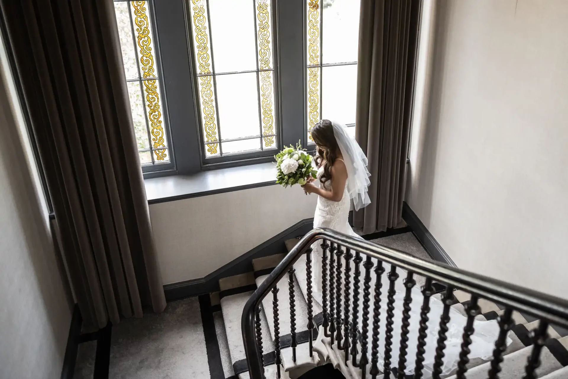 Carlowrie Castle bride descends the staircase in a white dress and veil, holding a bouquet. She is next to large windows with decorative glass panels.