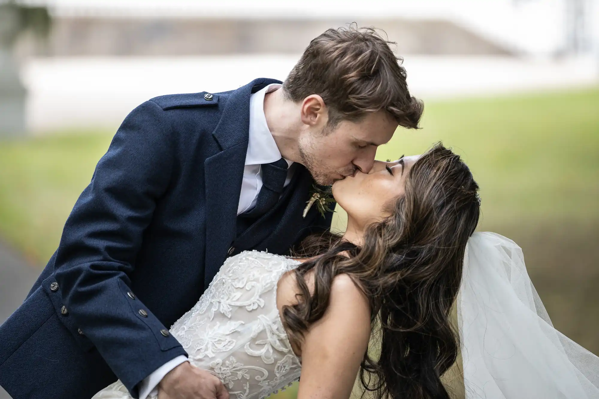 A bride and groom kiss outdoors. The groom is wearing a blue suit and the bride is in a white dress with a veil during their summer wedding at Carlowrie Castle.