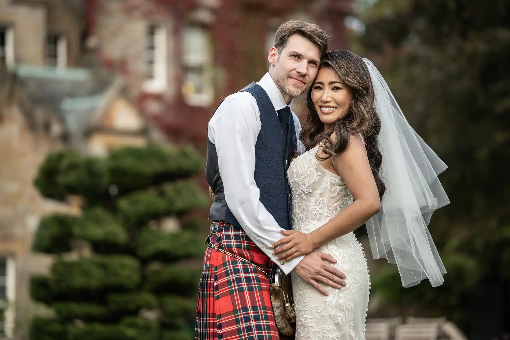 Carlowrie Castle wedding photography highlights by Edinburgh wedding photographers. Carlowrie Castle wedding photographer: A bride in a white dress and veil stands next to a groom wearing a kilt and vest, both smiling, in front of a lush garden backdrop.