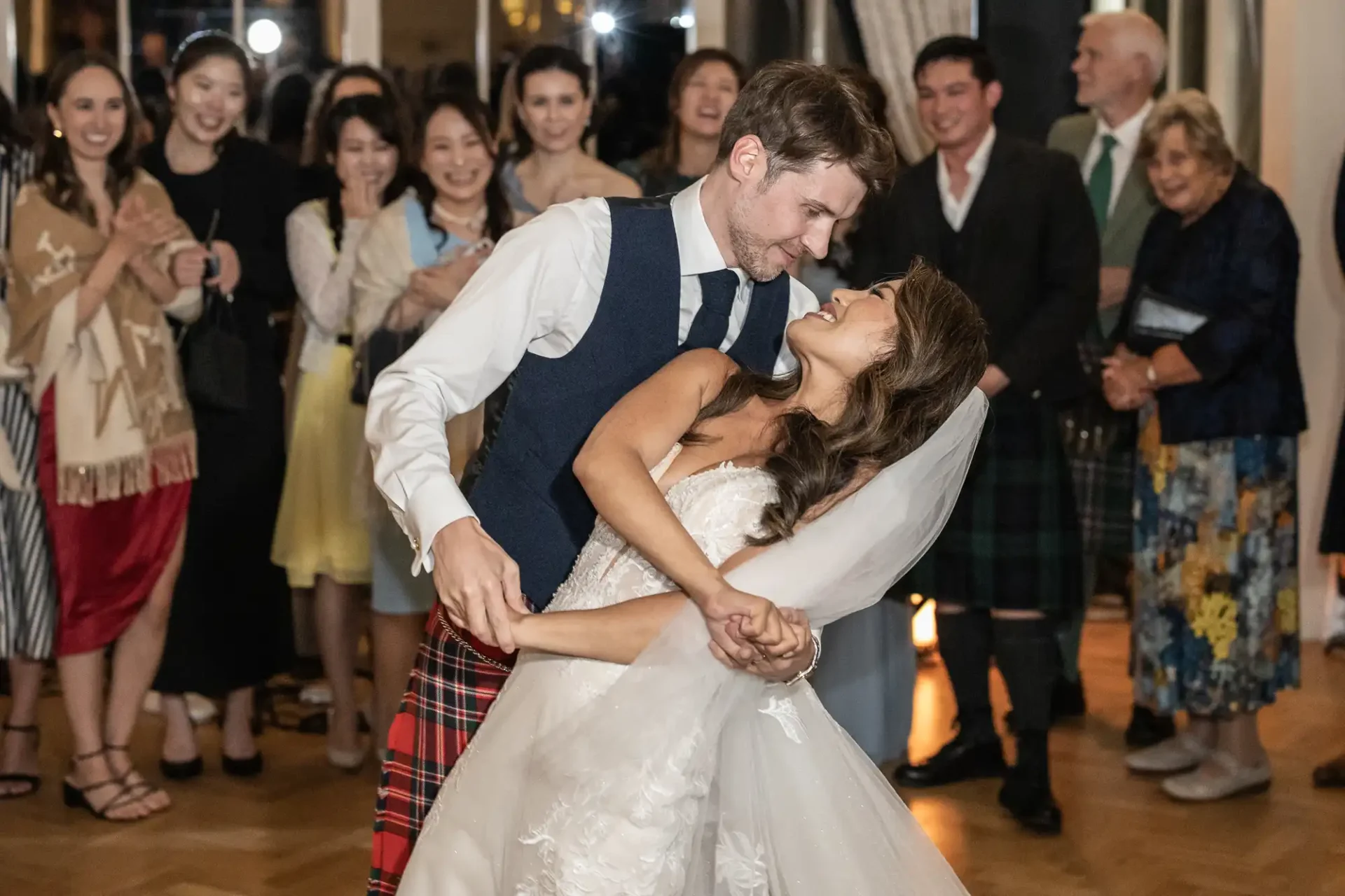 A couple in wedding attire dance joyfully, surrounded by clapping guests in a warmly lit Orangery wedding at Carlowrie Castle.