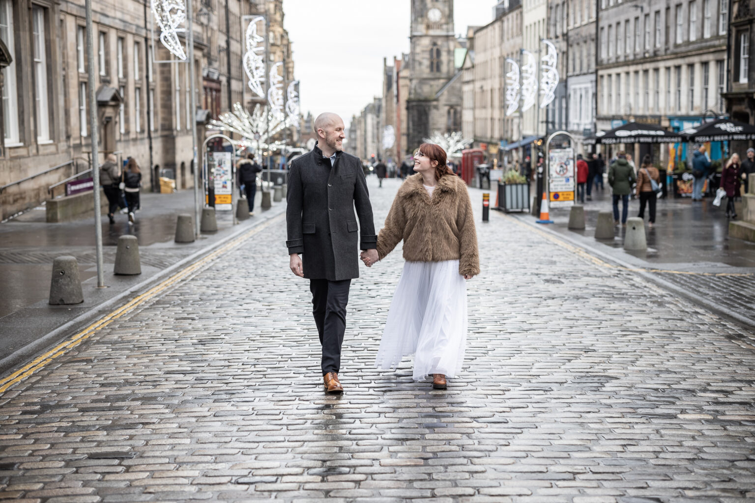 A couple walks hand in hand on a wet cobblestone street in a city. The man wears a dark coat and shoes, and the woman wears a brown jacket with a white dress. Festive lights hang above as people and shops line the pavements in the background.