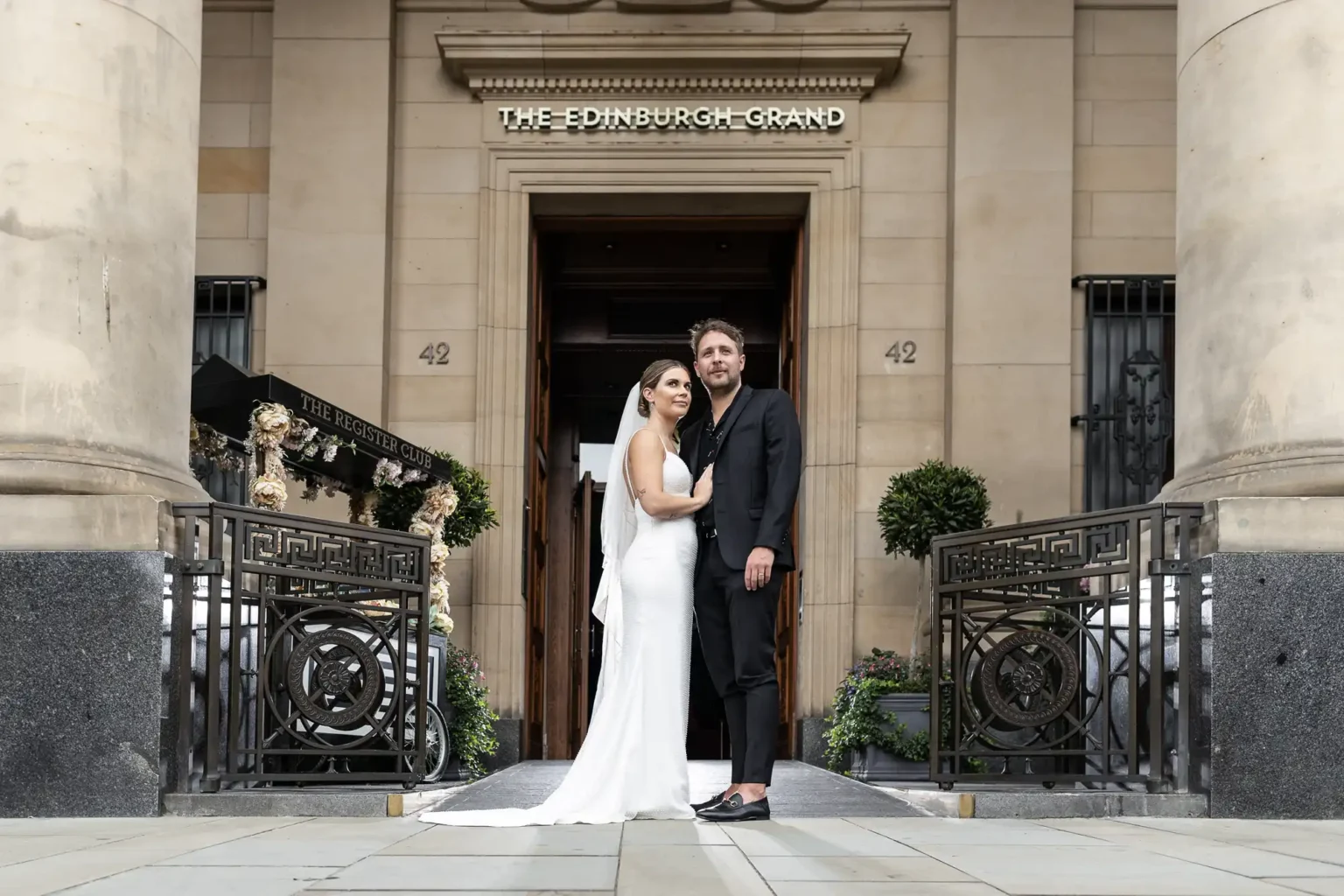 The Edinburgh Grand wedding photographer - luxury city centre wedding photography by Jonathon Fowler Photography. A bride in a white dress and veil stands beside a groom in a dark suit and sandals in front of The Edinburgh Grand entrance. They embrace, looking at the camera, surrounded by ornate railings, columns, and greenery on either side of the doorway.