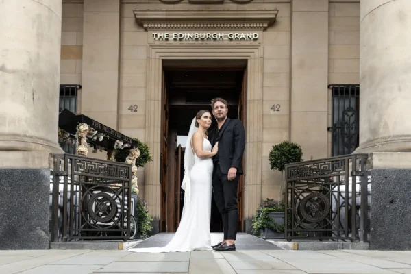 The Edinburgh Grand wedding photographer - luxury city centre wedding photography by Jonathon Fowler Photography. A bride in a white dress and veil stands beside a groom in a dark suit and sandals in front of The Edinburgh Grand entrance. They embrace, looking at the camera, surrounded by ornate railings, columns, and greenery on either side of the doorway.