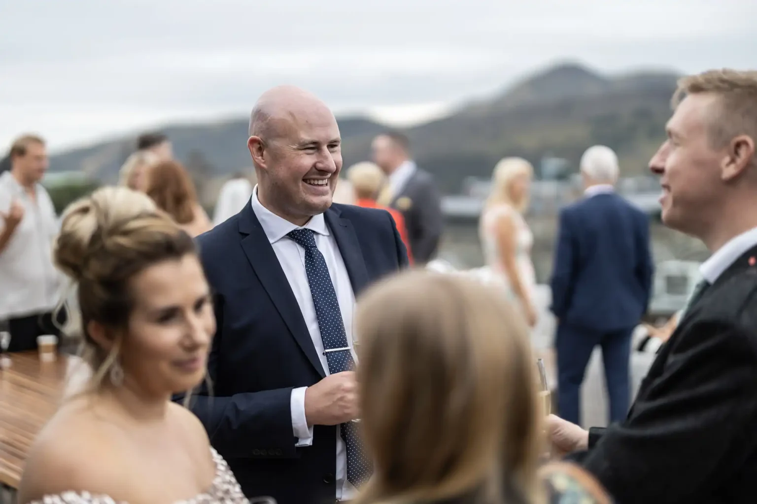A man in a suit laughs while talking with another man at an outdoor social event, with people in formal attire around them and hills in the background.