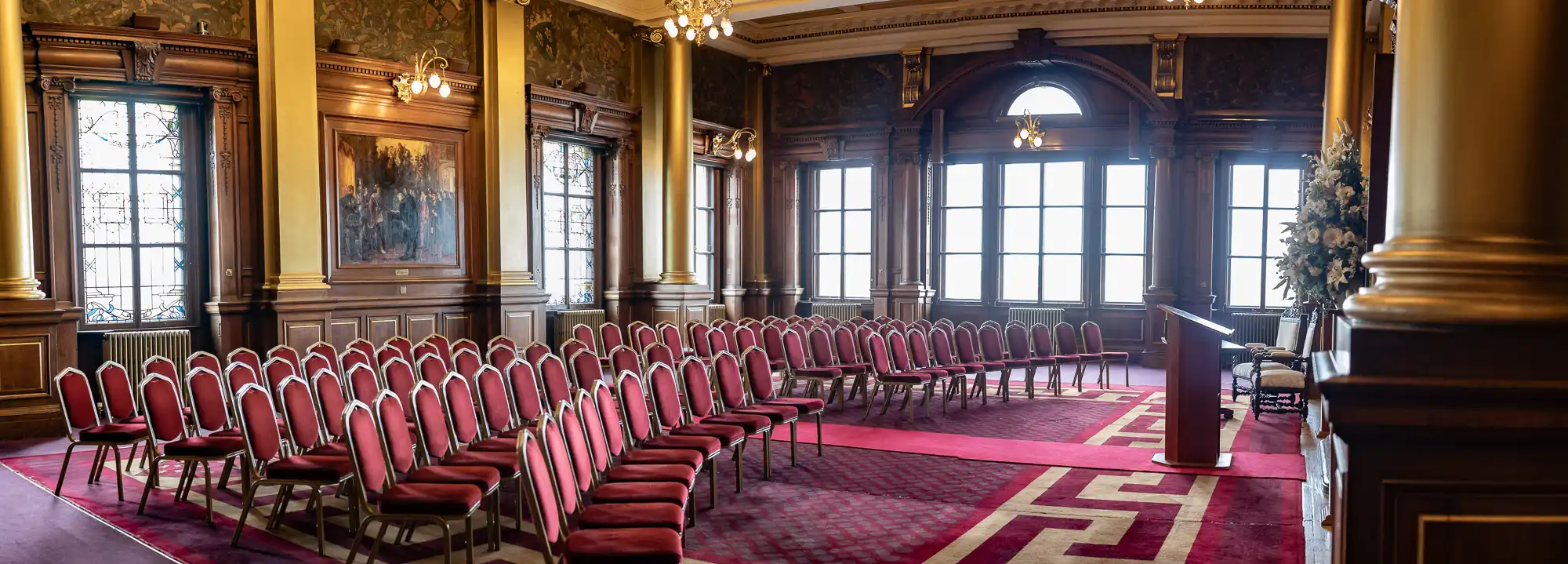 An elegant conference room with rows of red chairs, wooden paneling, large windows, and decorative chandeliers.