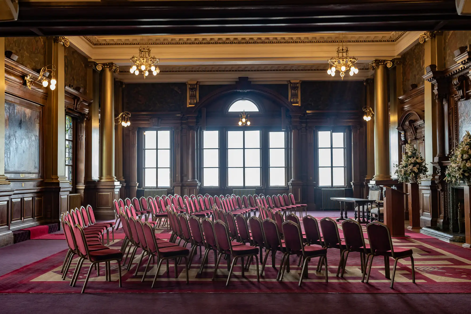 An elegant room with red chairs arranged in rows, under chandeliers, facing large windows and a wooden podium.
