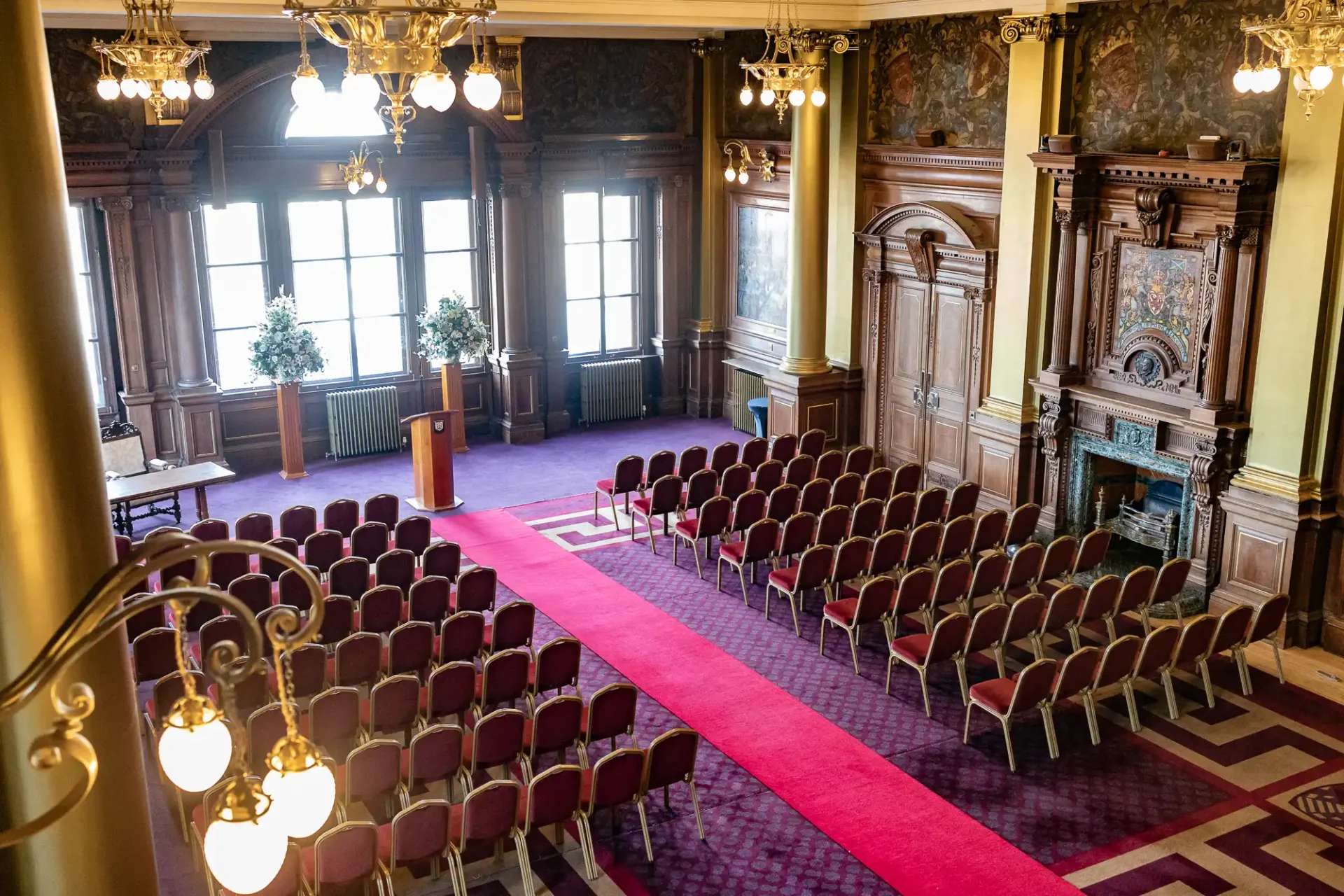Council Chamber wedding ceremony setup at Edinburgh City Chambers. An ornate room set up for an event with rows of red chairs facing a podium, surrounded by wooden paneling and large windows.
