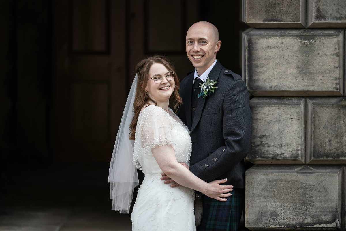 A bride in a white dress and veil stands with a groom in traditional attire. They are smiling and posing in front of a stone building.