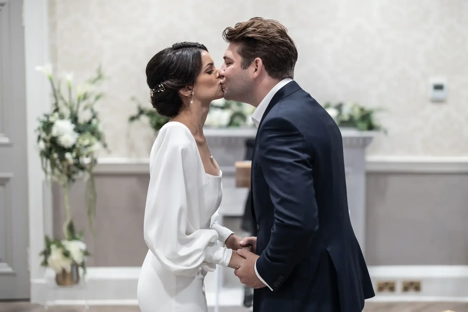 A bride and groom kissing at their wedding ceremony, with the bride in a white dress and the groom in a navy suit.