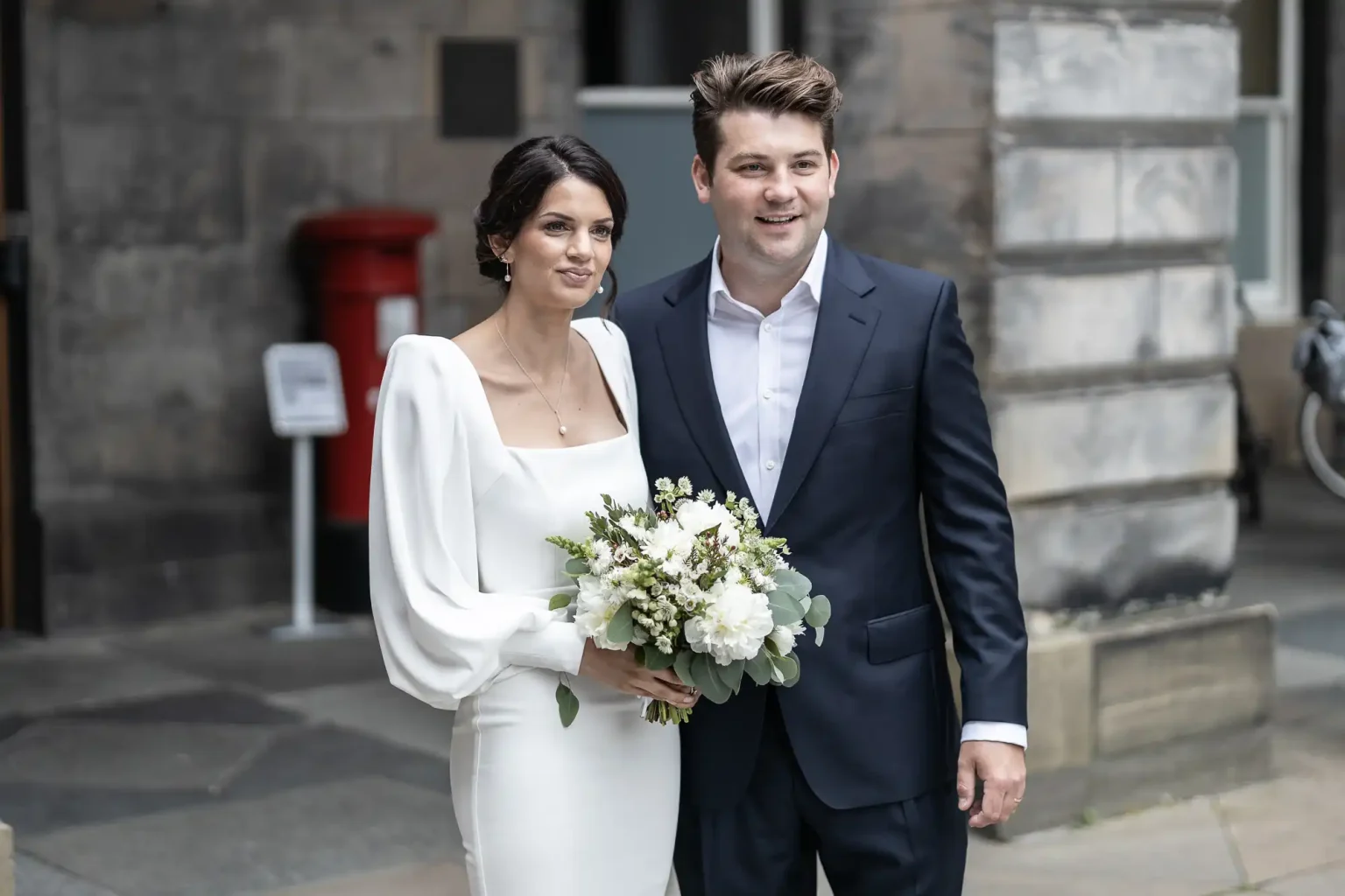 A newlywed couple walking on a city street, the bride holding a bouquet and both dressed in formal wedding attire.