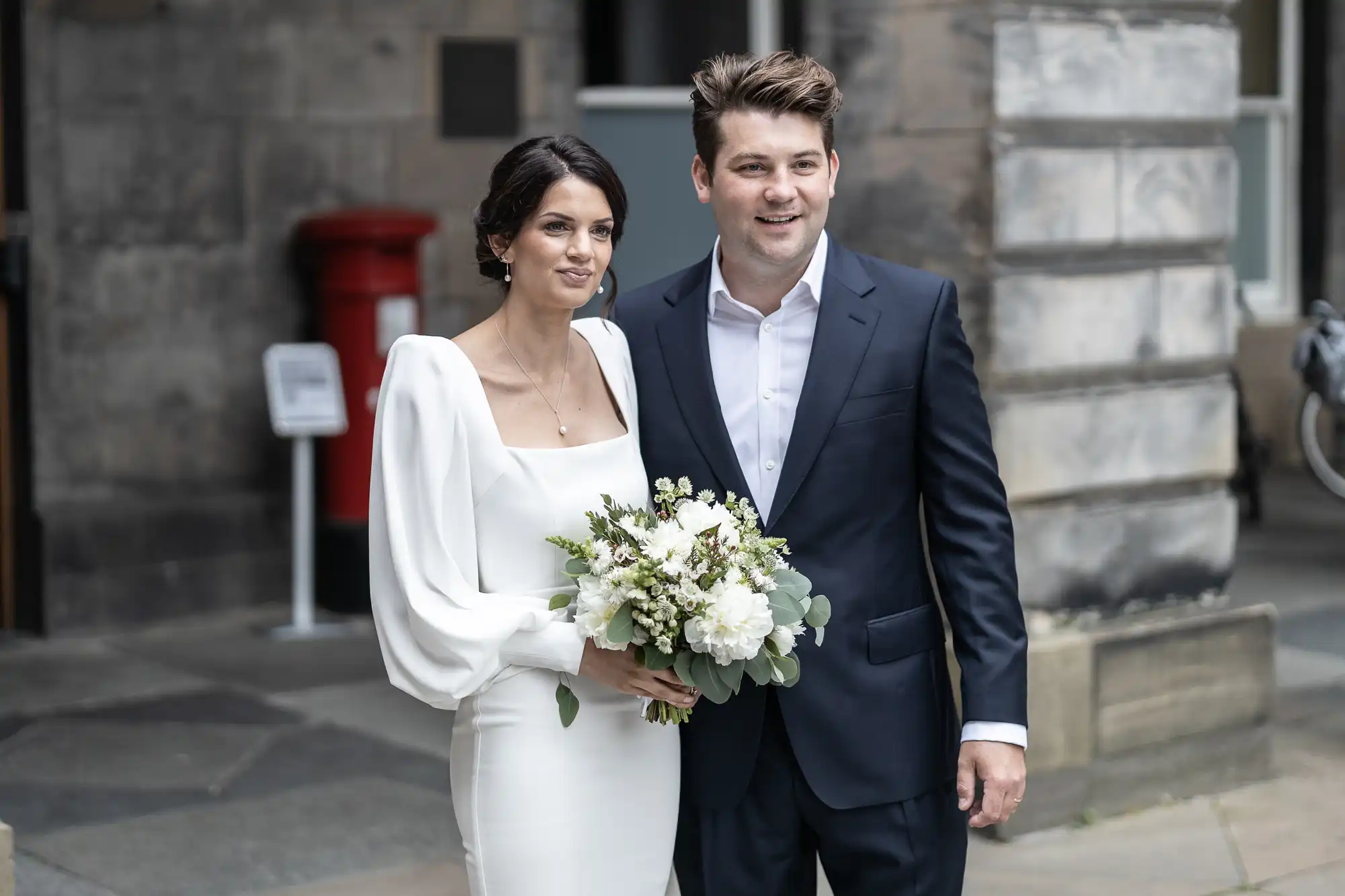 A newlywed couple walking on a city street, the bride holding a bouquet and both dressed in formal wedding attire.