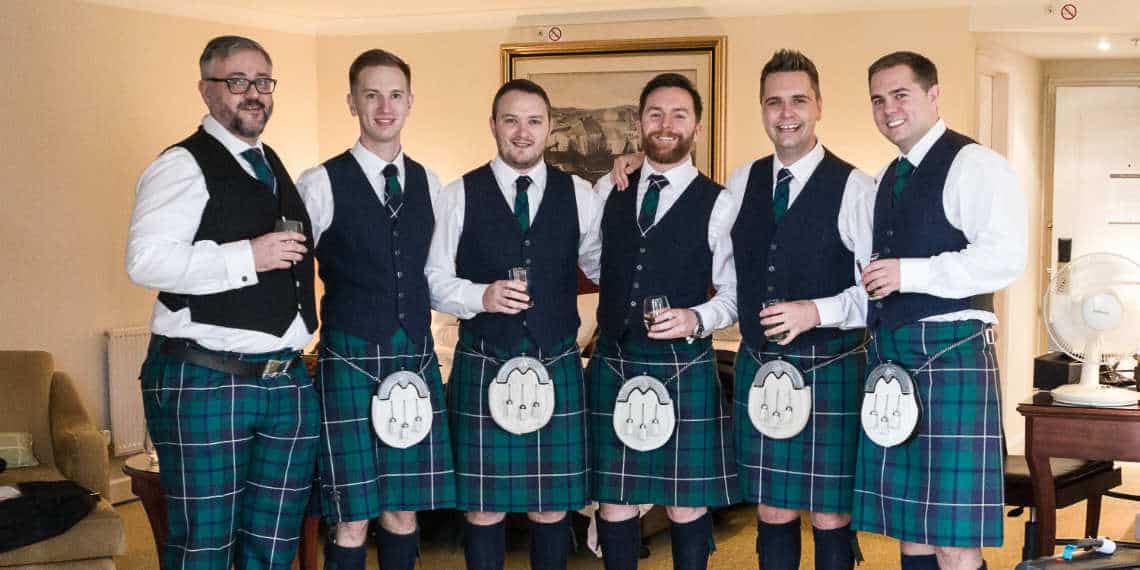 Six men in matching tartan kilts and waistcoats, holding drinks, smiling in a warmly lit room.