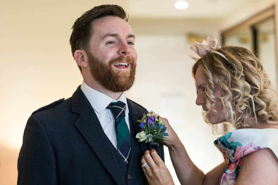 A woman adjusts a boutonniere on a smiling man's suit jacket at an indoor event, both dressed formally.