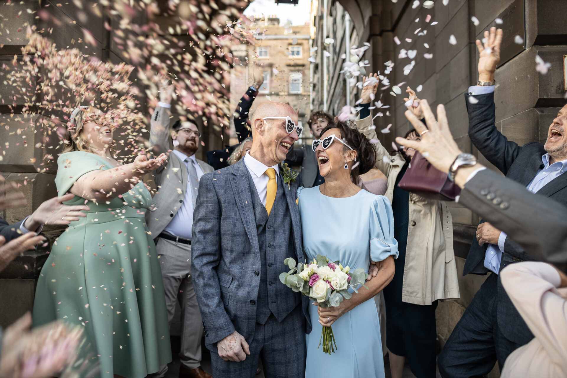 Newlyweds under confetti thrown on the Royal Mile outside Edinburgh City Chambers. The couple looks happy, and the woman holds a bouquet.