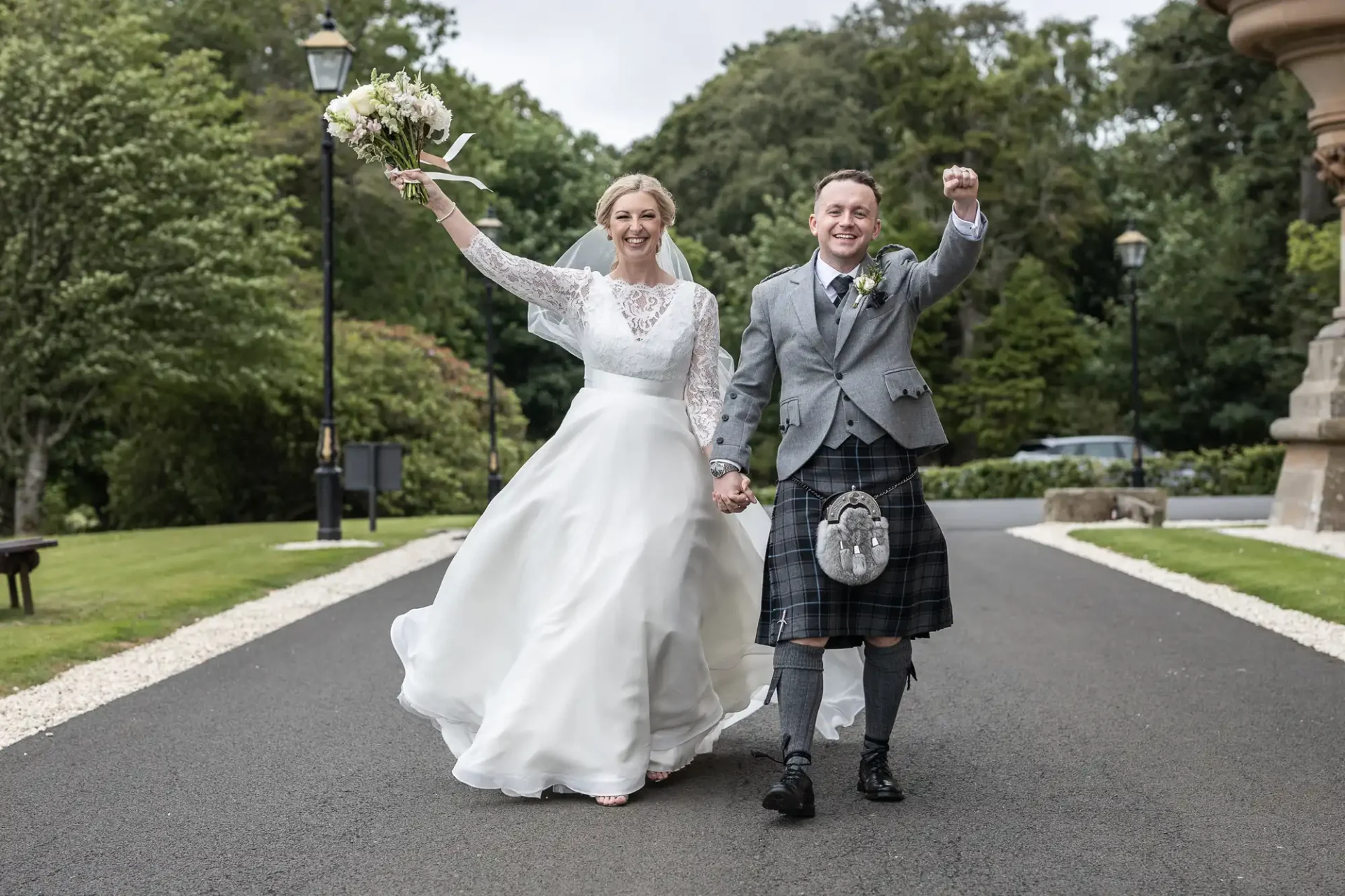 Cornhill Castle wedding photographer: A bride and groom walk hand-in-hand outdoors. The bride wears a white dress and holds a bouquet in the air. The groom wears a kilt and gray jacket, raising his fist in celebration. They are smiling.