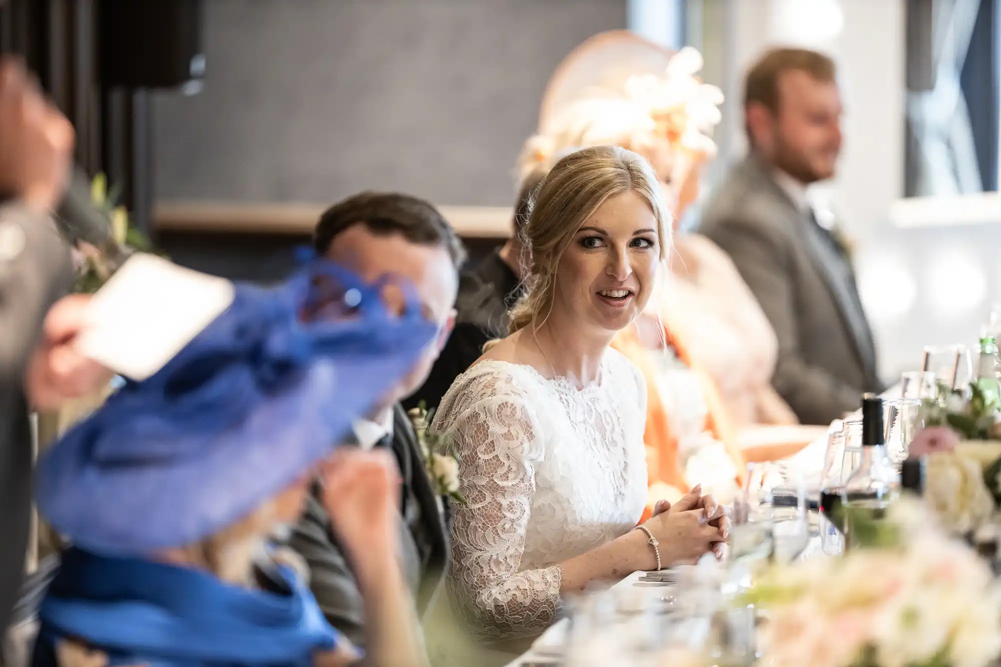 A woman in a white dress and veil is sitting at a table, smiling. Other people are seated next to her in formal attire. The setting appears to be a formal event or celebration.
