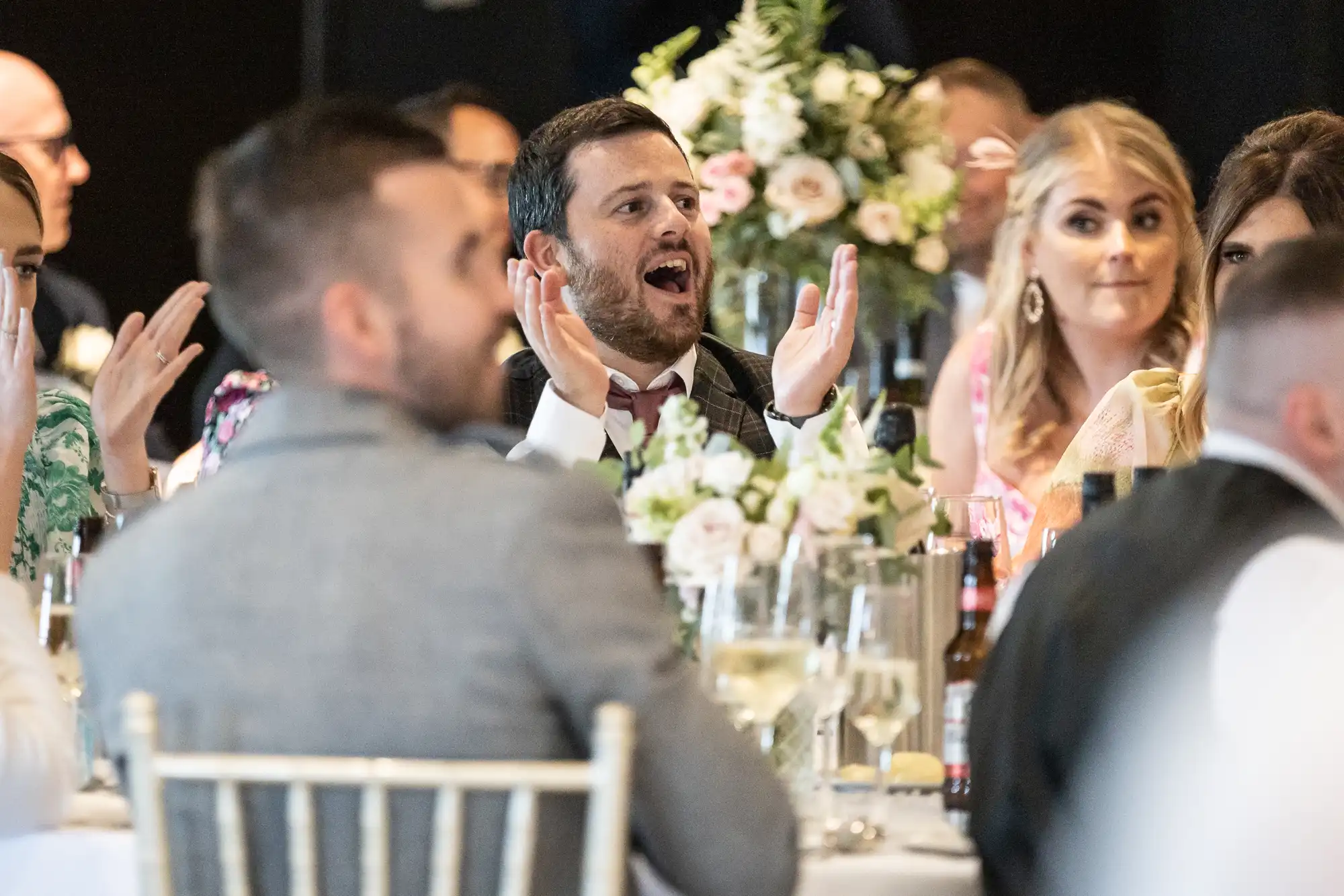 A group of people seated at a table with floral centerpieces, beverages, and dinnerware; a man in the center raises his hands mid-conversation.