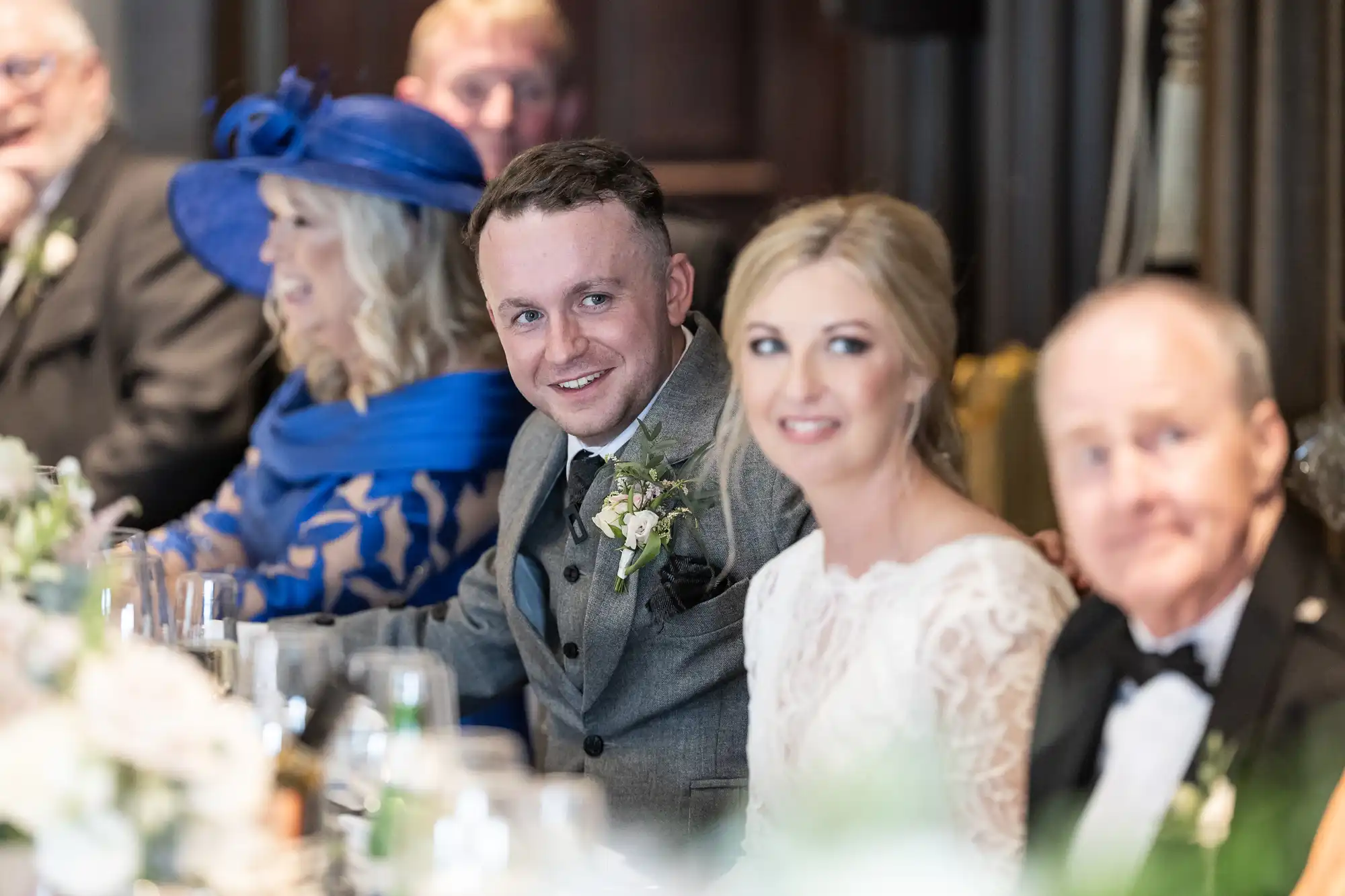 A bride and groom sit smiling at a wedding reception table with other guests. The bride wears a white dress, the groom a grey suit. A woman in a blue hat and dress is seated further along the table.