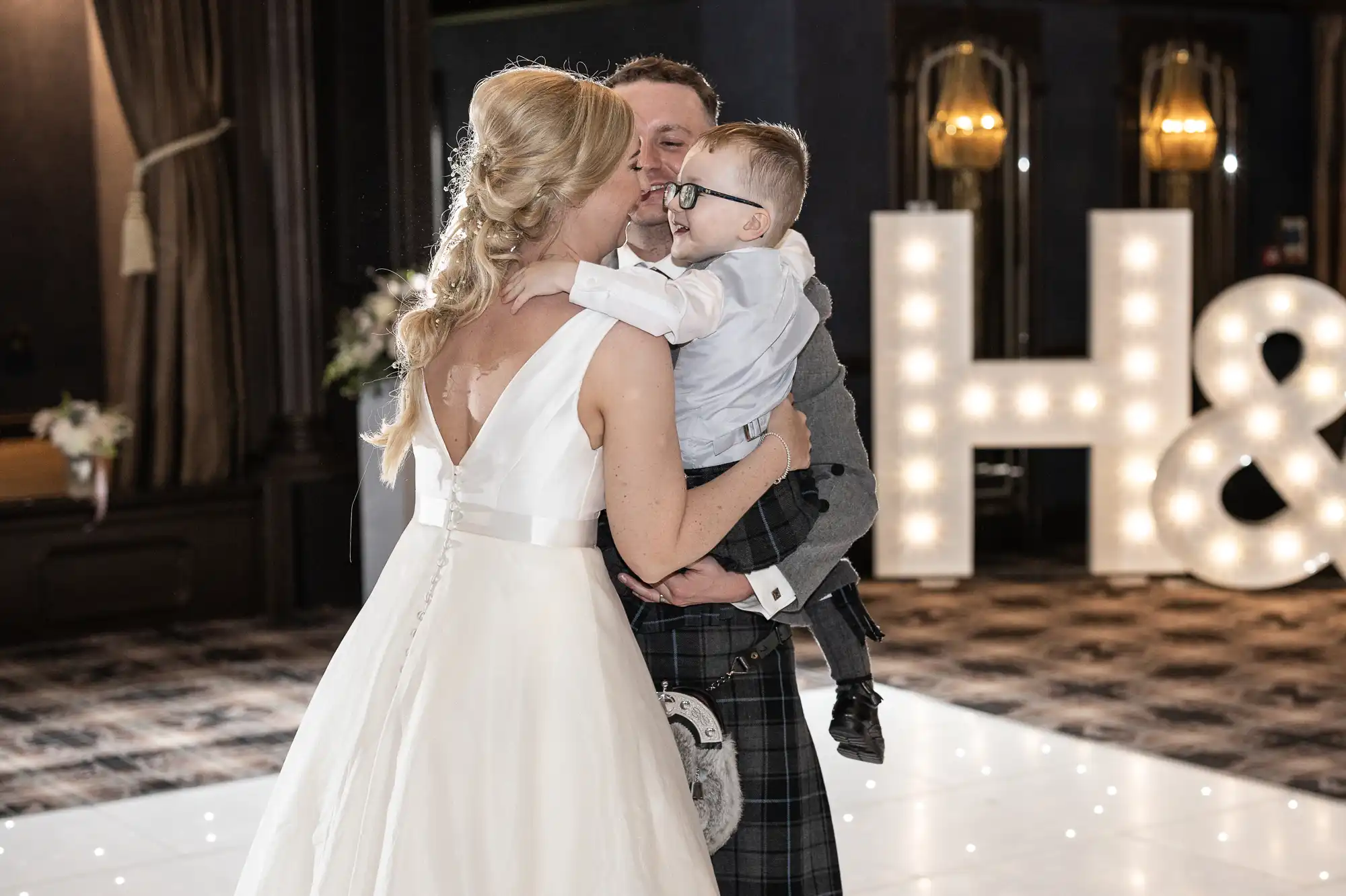 A couple in wedding attire shares a kiss while the groom holds a young boy. Large illuminated initials "H &" are in the background.