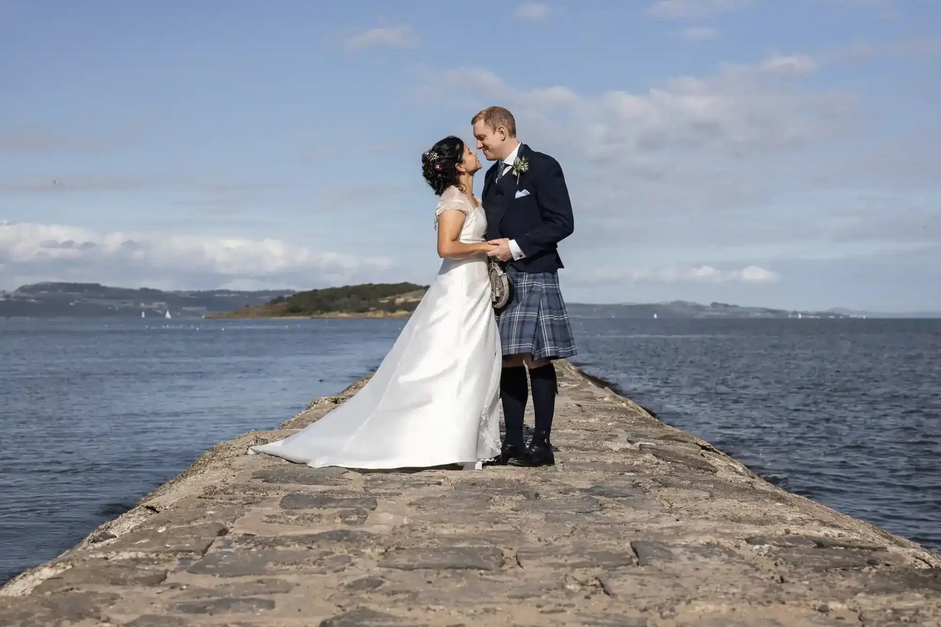 A newlywed couple stands on a stone pier by the water, the groom in a kilt and the bride in a white dress, facing each other under a blue sky.