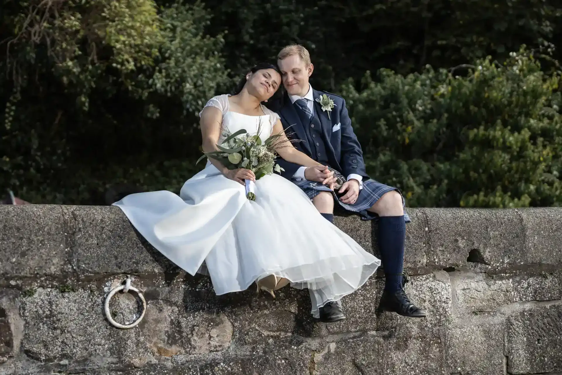 A bride in a white dress and a groom in a kilt sit on a stone wall, holding hands, with greenery in the background.