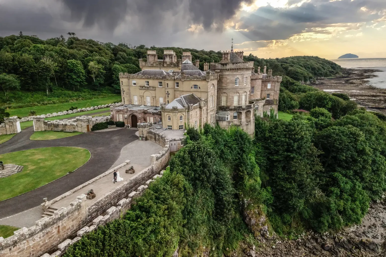 Drone wedding photography aerial view of a bride and groom standing beside large, historic stone Culzean Castle with multiple turrets, surrounded by lush greenery, located near a rocky coastline. The sky is partly cloudy with sunlight breaking through.