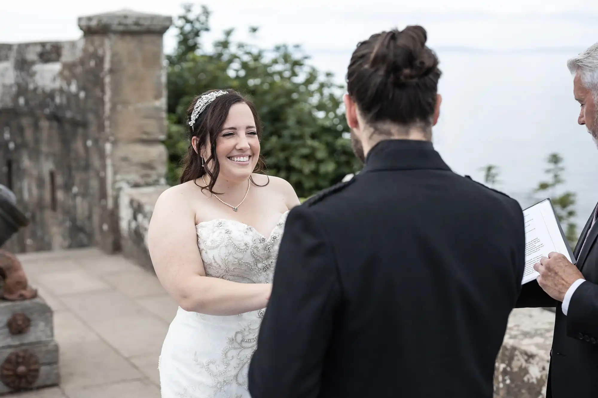 A bride and groom stand facing each other during their wedding ceremony, with an officiant reading from a document. Stone structures and greenery are visible in the background.