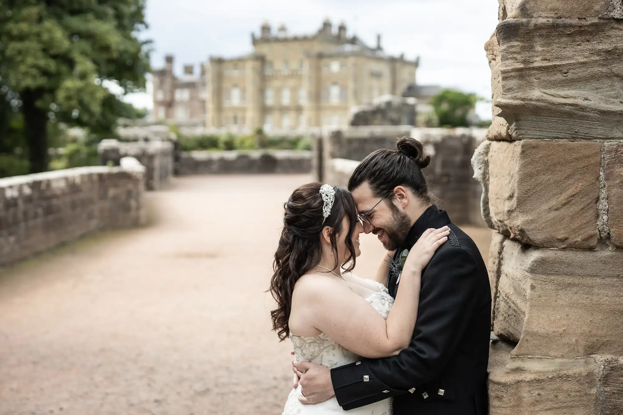 A couple in wedding attire embrace near a stone wall in an outdoor setting with a historic building in the background.