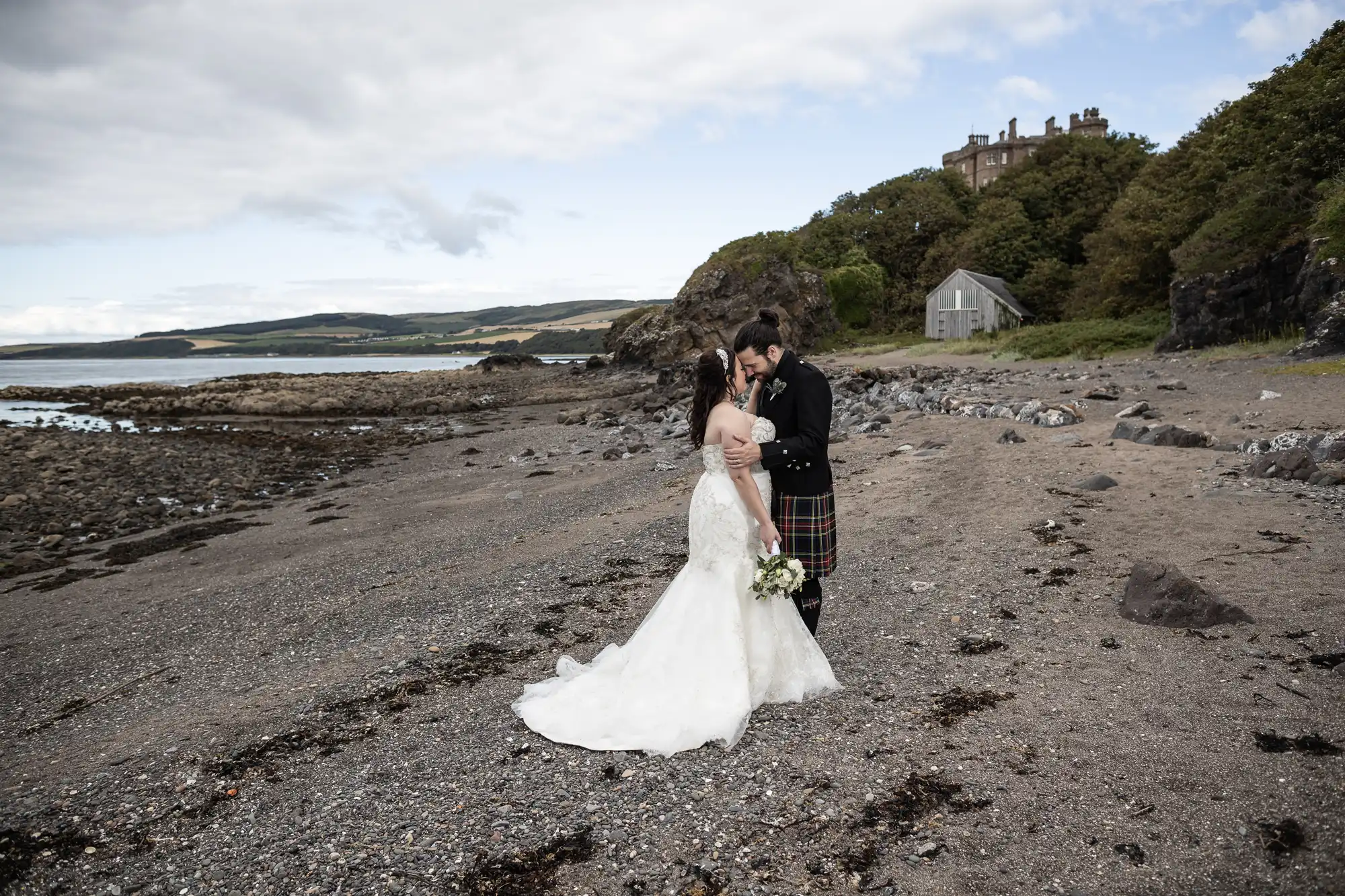 A bride in a white gown and a groom in a kilt embrace on a rocky beach with a house and trees in the background.