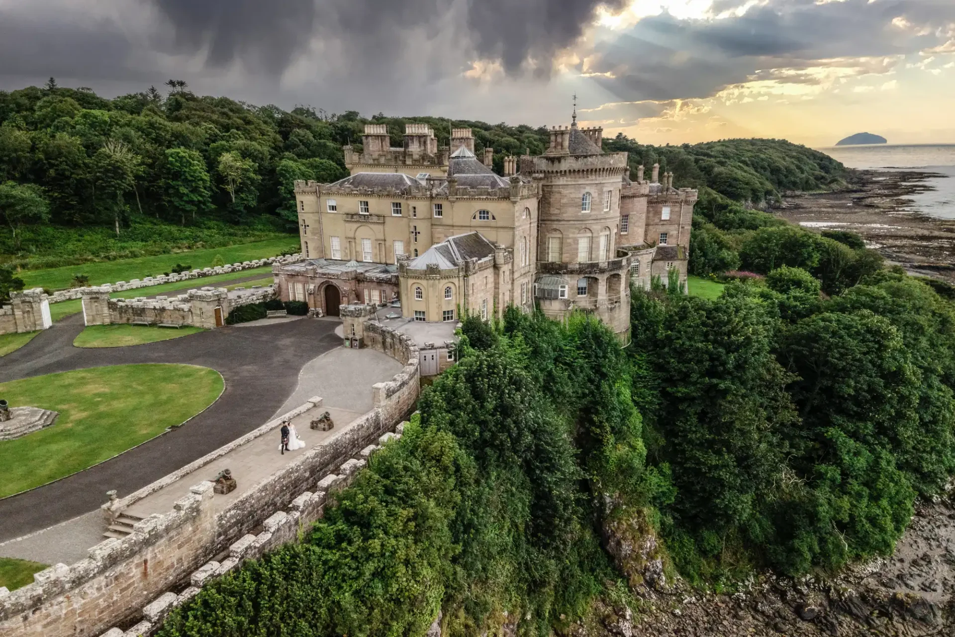 Aerial view of a large stone castle, Culzean Castle, situated on a cliff overlooking a coastline with lush greenery and a winding driveway.