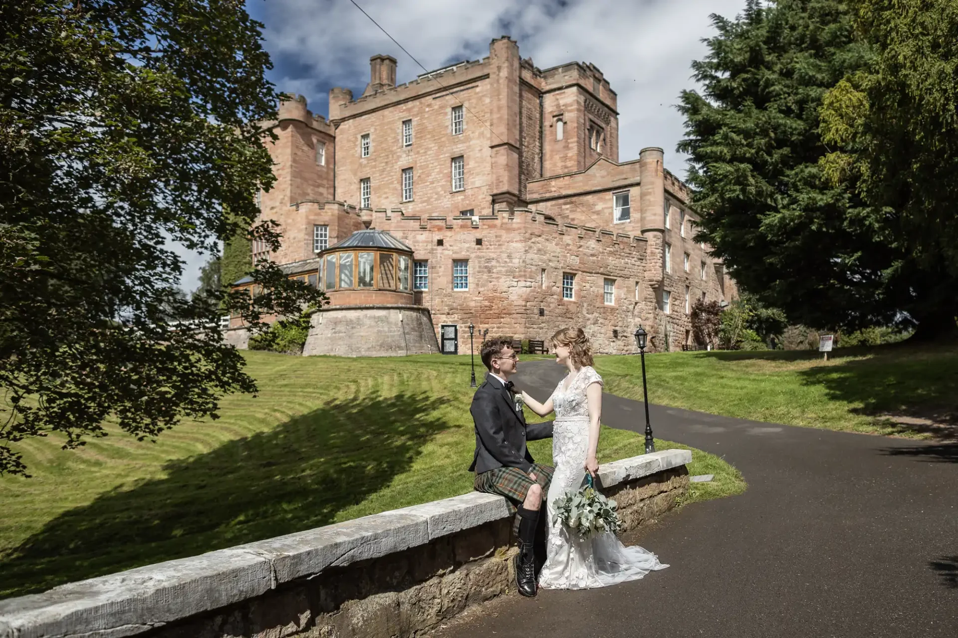 A couple stands beside a stone wall in front of historic Dalhousie Castle, one of the castle wedding venues Scotland, with the groom in a kilt and the bride in a white dress, holding a bouquet of flowers.