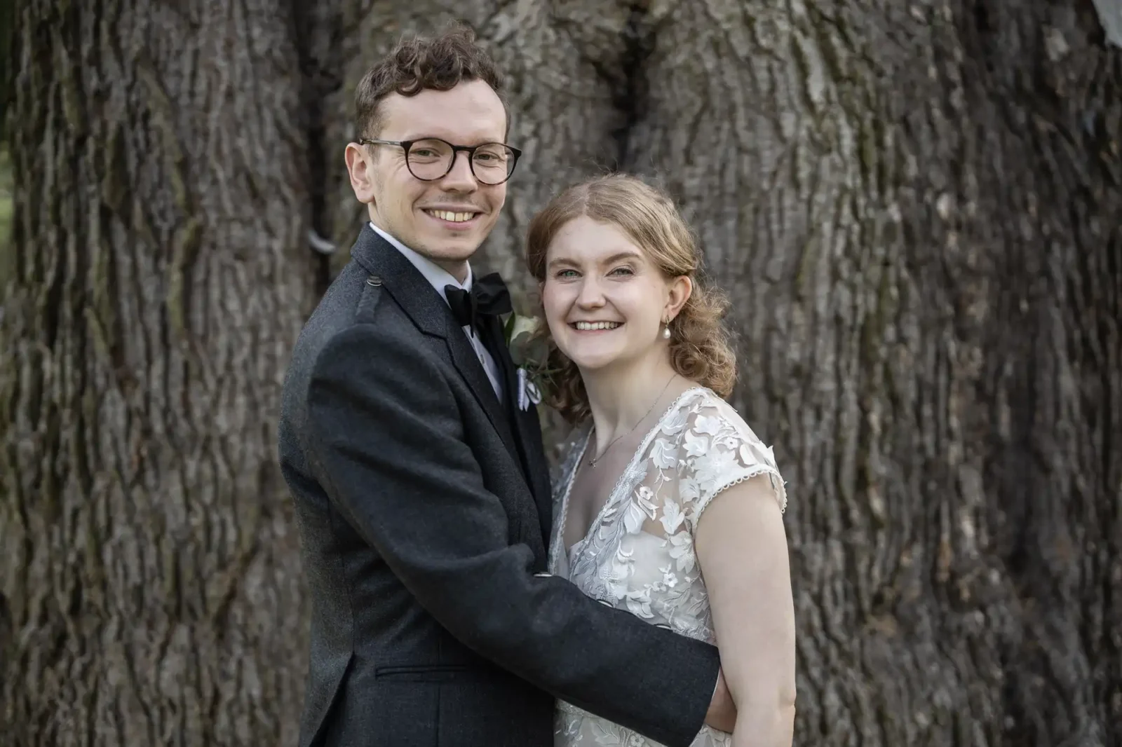 A smiling couple, the man in a dark suit and glasses and the woman in a white lace dress, stand embracing in front of a large tree.
