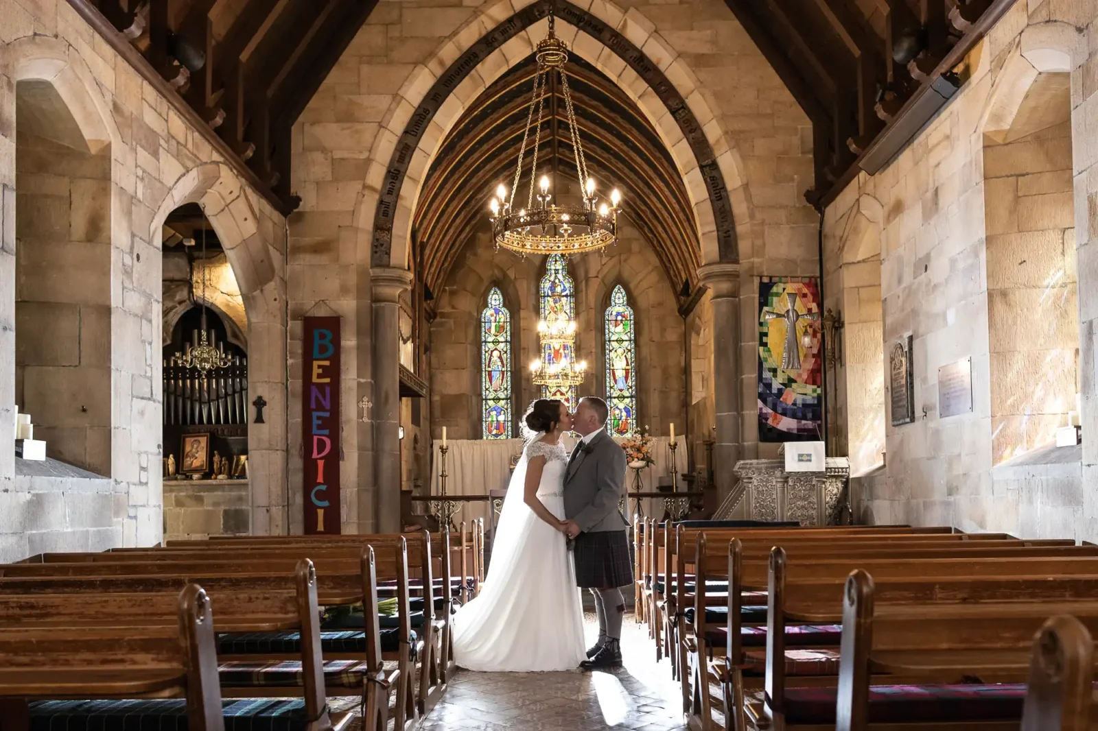 A couple, dressed in wedding attire, shares a kiss in the aisle of St Mary's Dalmahoy church with an arched ceiling, ornate stained glass windows, and wooden pews.