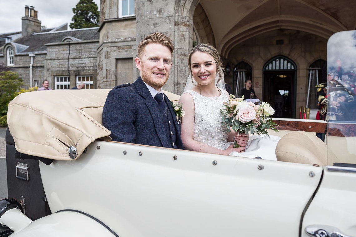 newlyweds sitting in the back of a convertible Beauford car at the entrance to the Dalmahoy Hotel