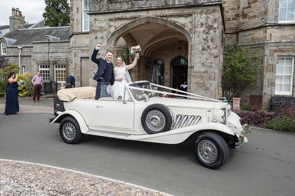 newlyweds standing in the back of a convertible Beauford car at the entrance to the Dalmahoy Hotel