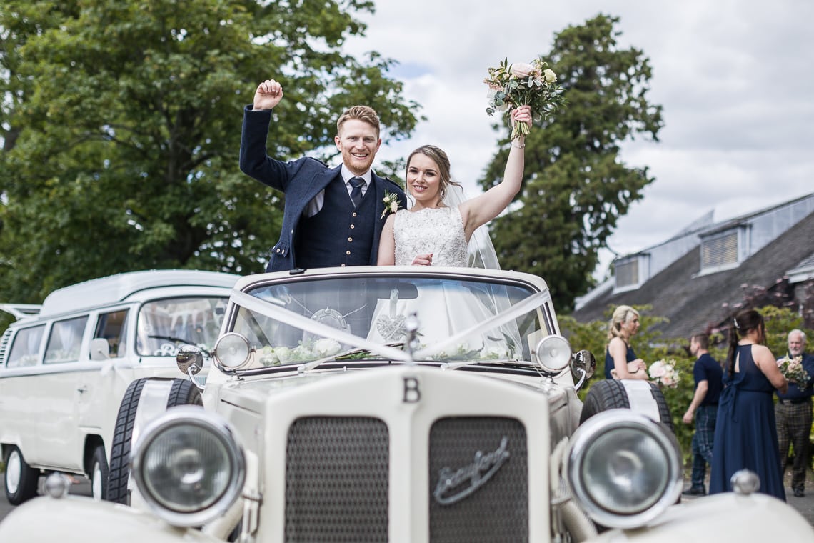newlyweds standing in the back of a convertible Beauford car at the entrance to the Dalmahoy Hotel